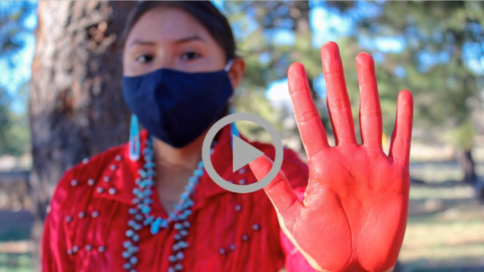 An Indigenous woman in traditional dress wearing a facemask holds her red-painted hand up to the camera. 