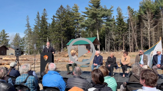 Deputy Secretary Beaudreau speaks to a crowd at an outdoor podium. Pine trees and a small building stand on a hill in the background.