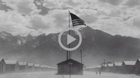Black and white image of an American flag waving in the breeze over the wooden structures of the Manzanar War Relocation Center.
