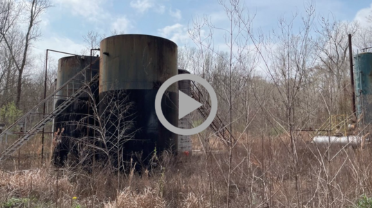 Several large and rusting fuel tanks sit abandoned and overgrown with weeds and small trees.