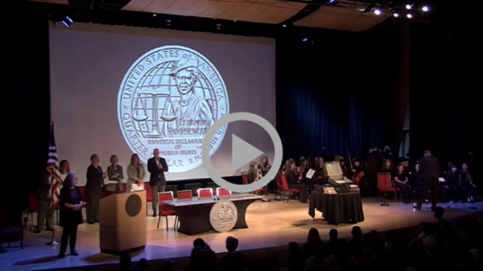 People gather on a stage with a large image of the new Eleanor Roosevelt quarter displayed on a screen behind them. 