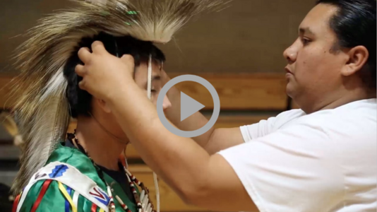 An Indigenous man adjusts a younger man’s ceremonial headdress. 