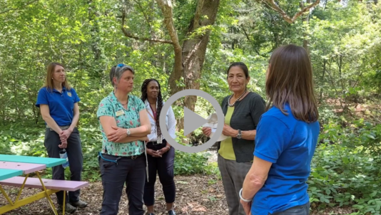 Secretary Haaland stands at the center of a group of women talking outside in a wooded, sunlit area.