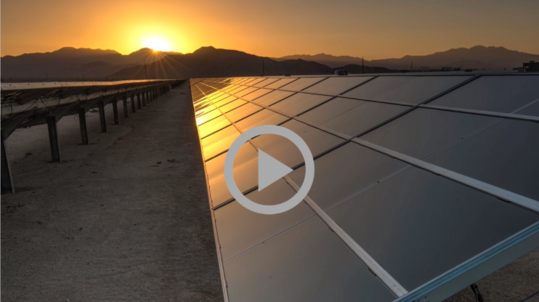 A solar energy array reflects the light of a sun setting over a desert mountain range.