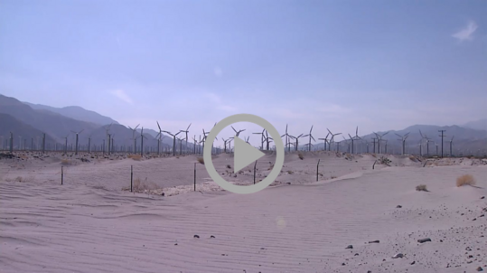 Dozens of wind turbines spin in the desert breeze in Southern California; mountains are visible in the distance.