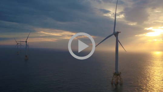 Wind turbines turn with the breeze at an ocean windfarm, with the sun setting through clouds. 