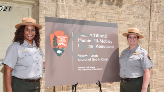 Two National Park Service Rangers stand beside a sign at the new Emmett Till and Mamie Till-Mobley National Monument. 