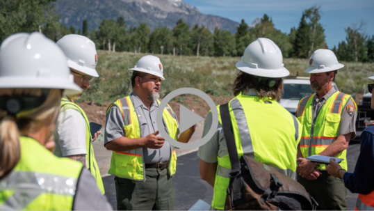 NPS Director Chuck Sams wearing a safety vest and hard hat speaks with NPS personnel at a Grand Teton National Park construction site.