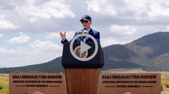Thumb: President Biden speaks from behind a podium with a cloudy sky, mountains and a grassy plain behind him.