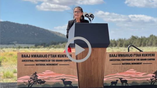 Secretary Haaland speaks from behind a podium with a cloudy sky, mountains and a grassy plain behind her.