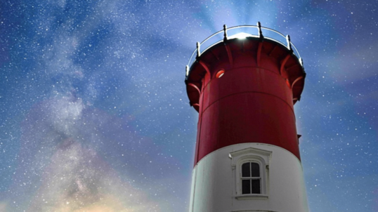 A red and white lighthouse shines bright underneath a night sky full of stars. The land where the lighthouse sits is surrounded by green trees.