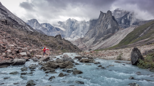 A person in a red jacket crosses a river in a valley with striking geologic Arrigetch Peaks rising thousands of feet into the clouds.