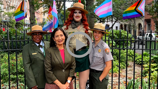 Secretary Haaland stands with Pattie Gonia and two other National Park Service representatives in front of a fence with rainbow flags. 