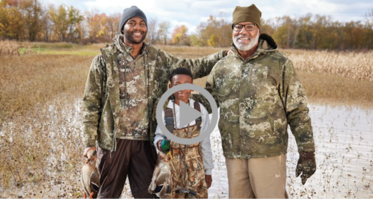 An older man, a younger man, and a boy pose with hunting trophies at a wetlands site in autumn. 