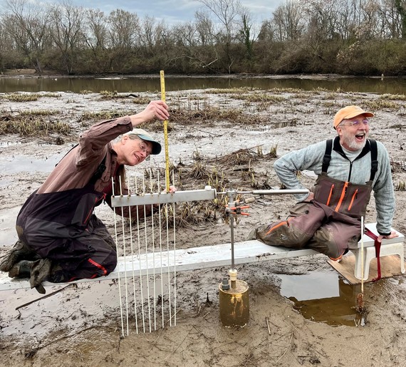 Sitting on metal bench over a muddy marsh, two scientists smile as they make measurements with a monitoring device. 