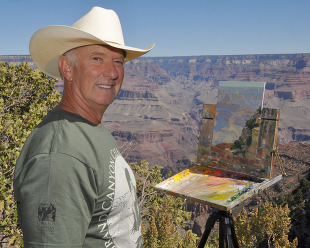 a smiling man next to a canvas with a scenic painting and a large canyon in the background