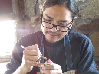 a man with glasses and long black hair crafting a small and intricate wooden toy