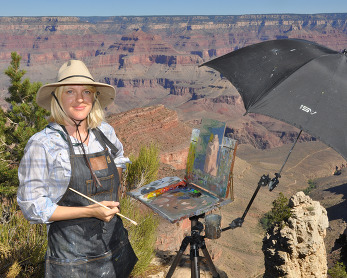 a smiling woman in front of a painting on a canvas next to the rim of a large canyon