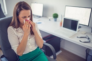 Picture of a woman sitting at a desk and covering her nose with a tissue. 
