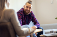 A young person wearing a blue and purple plaid button-up shirt sitting on a couch and speaking with a counselor in a professional setting.