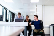 A diverse group of three people at desk in an office setting looking at a laptop together. One person uses a wheelchair.