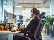 Photo of a woman using a wheelchair and headset, working at a desk with multiple monitors.