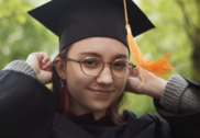 Young graduate in outdoor setting wearing glasses, a graduation gown, and mortarboard with orange tassel.