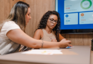 Two women sit next to each other at a work table in a teaching moment in front of a screen showing personal financial education material.