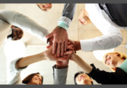 Six coworkers stand in a circle with hands stacked in a hand huddle, photographed from underneath.