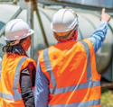 Two works on a job site in high visibility vests. One is looking where the other is pointing, as if providing assistance.