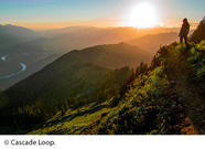 A hiker overlooks the sun setting over a river valley and mountain range. © Cascade Loop.