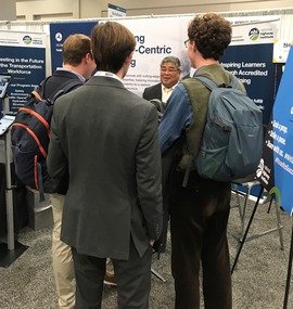 A smiling male NHI representative speaks with 3 young male attendees at NHI's booth at TRB's Annual Meeting.