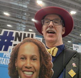 Male TRB attendee with white mustache and red fedora poses with a cardboard face cutout of NHI's director, a photo station prop.