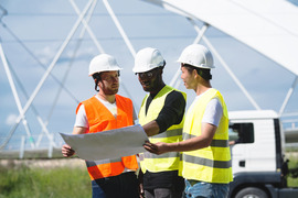 Three male construction workers or engineers in hard hats and construction vests holding plans with a bridge in the background