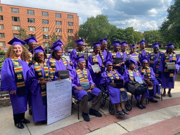 Eighteen people pose outside in graduation caps and gowns. There is an image of a Gallaudet Proclamation on poster board.