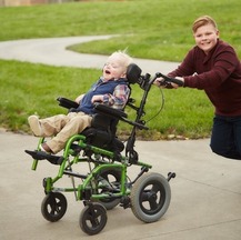 A boy is pushing his younger brother who is deafblind in a wheelchair. They are outside and both are smiling broadly.