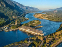 A photo of a water power station on a blue river surrounded by forested hills.
