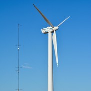 A three-bladed wind turbine installed on the ground with a meteorological tower and blue sky in the background.