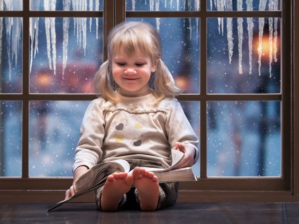 A young girl reading a book in front of an icy window.