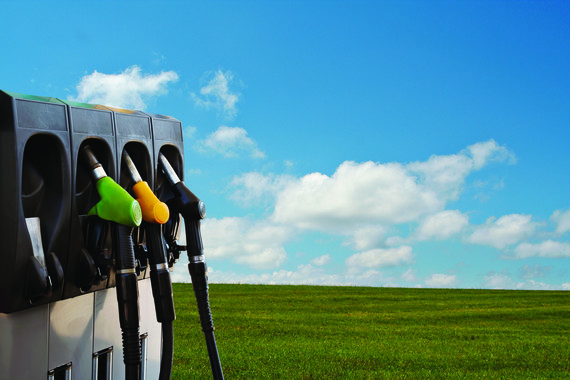 Photo of a fuel pump with three different colored handles in the foreground, with a field and blue sky in the background. 