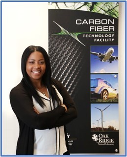 A vertical photograph of a woman standing in front of a sign that is roughly the same size as her. The woman has dark skin and long, dark hair. 