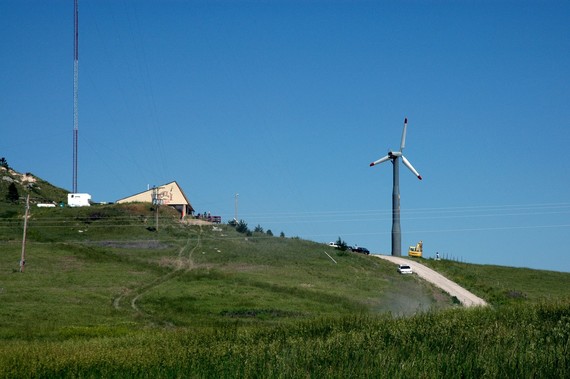 A tractor and cars surround a lone wind turbine near a radio station building in a rural, grassy area.