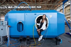 A woman sits in the porthole opening of a car-sized device.