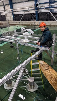 A student wearing a hard hat and waterproof attire sits on a structural beam above a tank of water. 