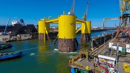 Three connected columns being towed by a boat from a water channel next to harborside cranes.