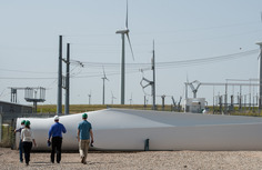 People in hard hats walking toward a wind turbine laying on a field with operating turbines and power lines in the distance.