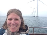 Woman smiles for camera in front of line of water-bound wind turbines. 