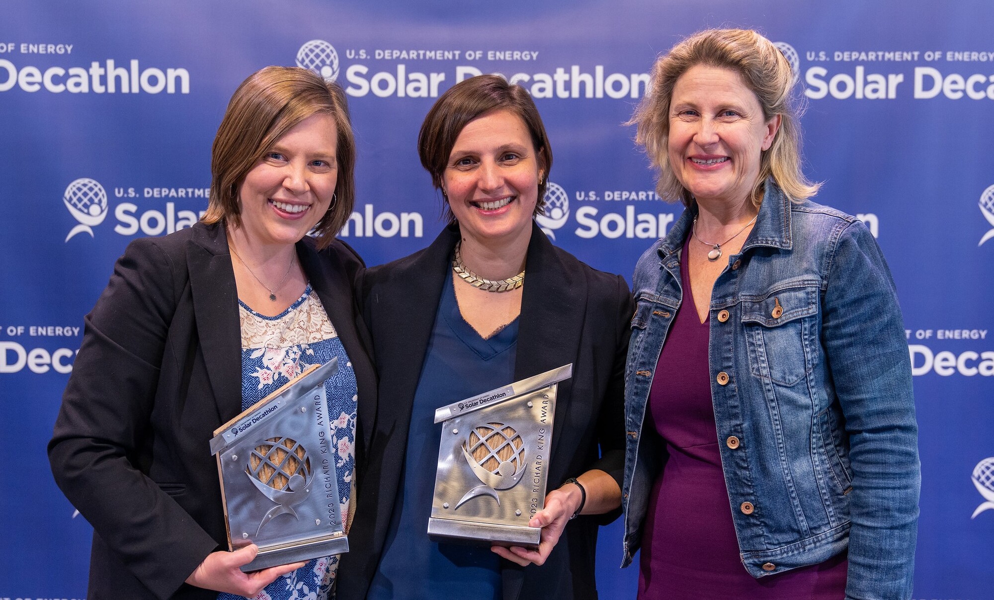 Three women smiling and looking at the camera, holding awards, against a backdrop with the Solar Decathlon logo.