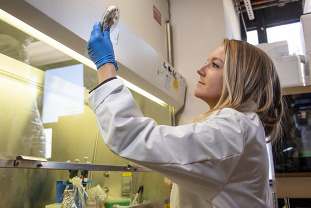 A person in lab coat and gloves holds up a sample to the light