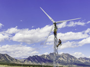 Two people climb a wind turbine with mountains in the background. 